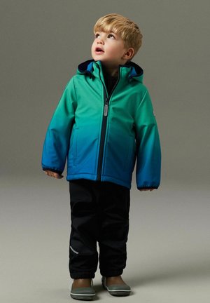 Fiatal gyermek zöld-kék átmenetes dzsekiben, fekete nadrágban és szürke cipőben, állva, felfelé nézve, egyszínű háttér előtt.