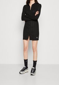 Robe noire à manches longues avec boutonnage sur le devant, col et une fente frontale. Présente un motif texturé et un logo CK en bas à droite. Portée avec des baskets et des chaussettes.
