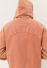 Hoodie u izblijedjelom koraljnom tonu s mekanom teksturom. "LES DEUX" je izvezena u zakrivljenom luku na leđima svijetlom niti.