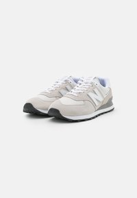 Para beżowo-białych sneakersów New Balance z białymi sznurowadłami i czarnymi gumowymi podeszwami na białym tle.