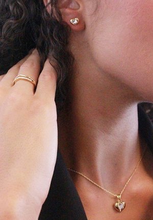 Collier en pendentif en or en forme de cœur, petites boucles d'oreilles en or avec pierre claire, bagues superposées sur la main, peau texturée et cheveux ondulés visibles.