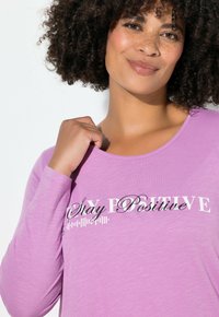 Laventelinvärinen pitkähihainen paita, jossa pyöreä pääntie ja "Stay Positive" -teksti; pehmeä, kevyt kangas, jossa sileä pinta.