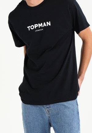 Personne portant un t-shirt noir "TOPMAN LONDON" et un jean bleu, les mains derrière le dos sur un fond blanc.