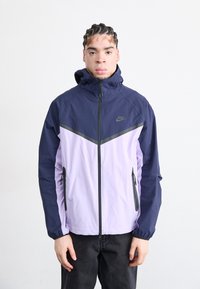 Veste coupe-vent navy et lilas clair avec fermeture éclair à l'avant, capuche et poches latérales ; présente un motif en chevrons et des accents noirs.