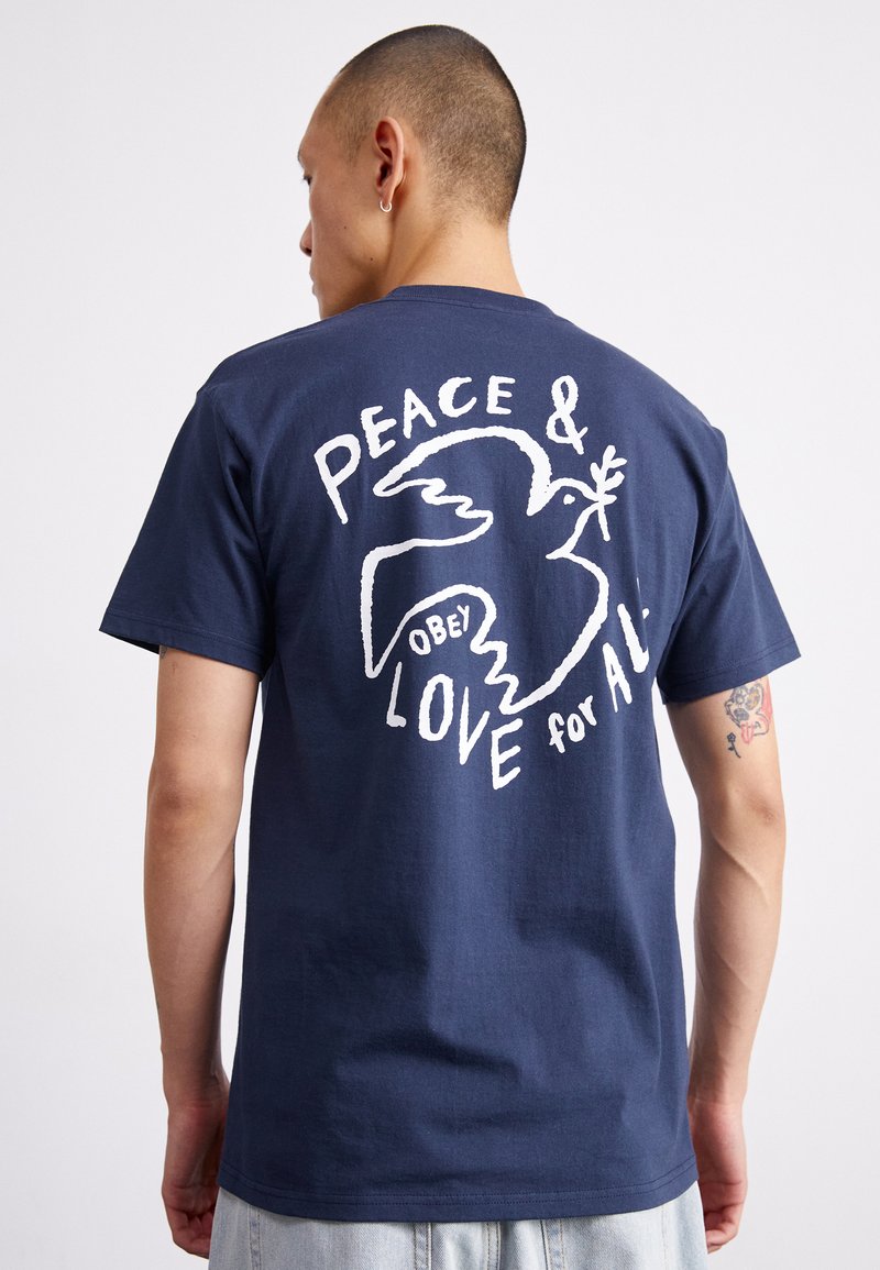 Camiseta de algodão azul marinho com uma impressão gráfica branca de uma pomba com os textos "PAZ & AMOR para TODOS" e "OBEDECE" nas costas. Mangas curtas.