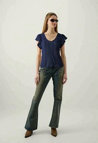 Mulher vestindo uma blusa azul-marinho de manga curta com mangas de babado, jeans verdes de boca de sino, botas marrons de bico fino, óculos de sol e brincos de argola, de pé em um fundo simples.