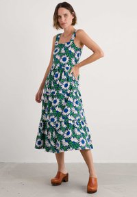 Femeie în picioare purtând o rochie midi fără mâneci, în nuanțe de albastru, alb și verde, cu flori și buzunare, împreună cu clogs din lemn maro.