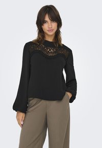 Top noir à manches longues avec un panneau en dentelle au design ondulé sur la poitrine. Texture lisse, col montant, associé à un pantalon marron clair.