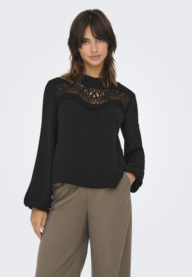 Top noir à manches longues avec un panneau en dentelle au design ondulé sur la poitrine. Texture lisse, col montant, associé à un pantalon marron clair.