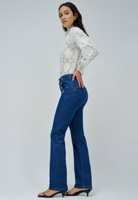 Jeans de tiro alto de color azul con piernas acampanadas, combinados con una blusa blanca de encaje de manga larga con patrones intrincados. Sandalias negras de tacón.