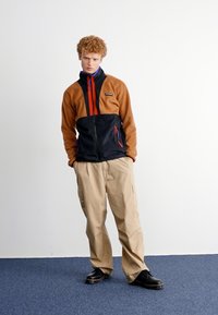 Fleece zip-up jakke med brun topp og svart nederste del, med røde og blå accenter. Beige cargo bukser og svarte sko fullfører antrekket.