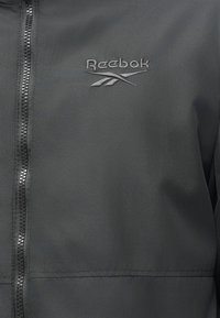 Czarna kurtka zapinana na zamek wykonana z gładkiego, lekkiego materiału. Posiada logo Reebok w ciemnym haftowanym znaku, z zamkiem z przodu i bez dodatkowych akcentów.