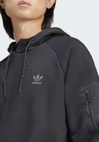 Svart hoodie med en logga på bröstet, texturerad tyg, snören i detaljerna och en ficka med dragkedja på ärmen.