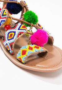 Sandale en cuir avec des brides ornées de perles colorées, des pompons rouges et verts, et des décorations en forme de breloques dorées sur fond blanc.