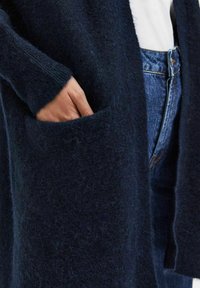 Cardigan blu navy con una texture morbida e lanosa, apertura frontale, maniche lunghe e una grande tasca laterale. Indossato sopra una maglietta bianca e jeans di denim.