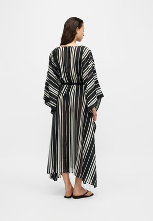 Femme debout, dos tourné, portant une longue robe rayée noire et blanche à manches larges et des sandales noires.