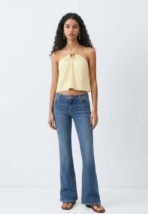 Femme aux cheveux bouclés porte un haut à col licou jaune, un jean flare bleu, des boucles d'oreilles et des sandales, se tenant devant un fond blanc uni.