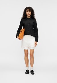 Μαύρο πουκάμισο με μακριά μανίκια και κουμπιά, λευκά σορτς με διακοσμητικά τρυπήματα, πορτοκαλί τσάντα και μαύρα μοκασίνια με υφασμένα πέλματα.