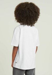 Hvid bomulds t-shirt med korte ærmer, lige pasform, med et lille logo-tagg nær sømmen. Bagudskuet, model iført grå bukser.