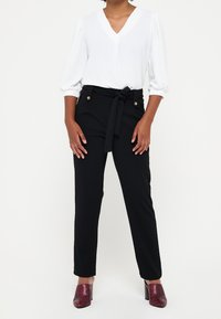 Bílá blůza s výstřihem do V a nabíranými rukávy, spárovaná s černými kalhotami s vysokým pasem, které mají opasek na zavazování a zlaté knoflíkové akcenty. Červené vzorované lodičky.