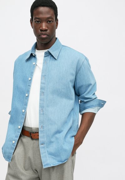 Homem vestindo uma camisa de denim azul claro sobre uma t-shirt branca, calças cinzentas e um cinto castanho, de pé com uma mão no bolso contra um fundo branco.