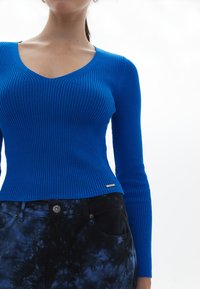 Maglione a V, con maniche lunghe, in un vibrante blu a costine, abbinato a pantaloni neri con effetto tie-dye, dotati di tasche e dettagli in metallo sui bottoni.