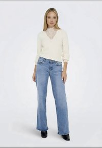 Pull en maille blanche avec un détail en dentelle au niveau du col, associé à un jean flare bleu clair. Modèle portant des bottes à talons noires.