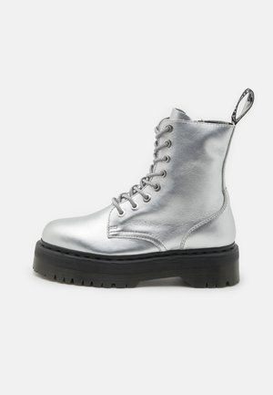 Botte de combat montante en cuir métallisé argenté avec semelle noire épaisse et talon à boucle d'extraction sur fond blanc.