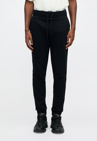 Black sweatpants with a drawstring waistband and logo detailing. Featuring a tapered fit and ribbed cuffs. Paired with black sneakers.