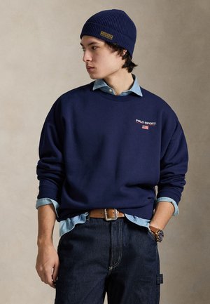 Marineblå sweatshirt med "POLO SPORT" logo, lag-på-lag over en lyseblå denimskjorte, kombineret med mørke jeans og en matchende strikket beanie.