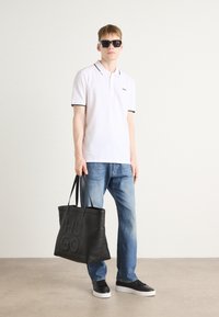 Polo bianco con cuciture nere, jeans blu e sneakers nere. In mano una grande borsa a tracolla nera con logo "HUGO" in rilievo.
