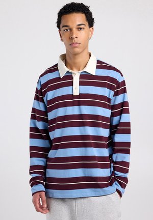 Young man with curly hair wearing a long-sleeve rugby shirt with blue and maroon horizontal stripes and light-colored collar, standing against neutral background.