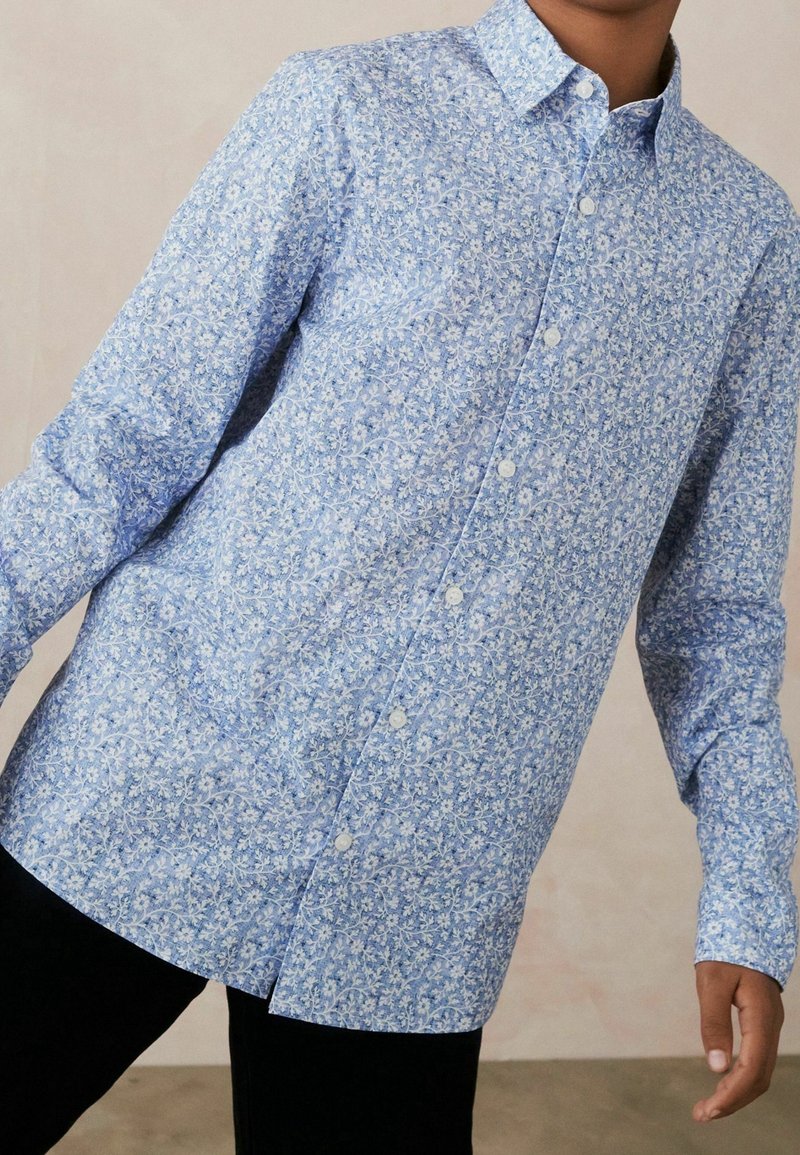Personne portant une chemise bleu clair à manches longues boutonnée avec des motifs floraux blancs et un pantalon noir sur un fond neutre.