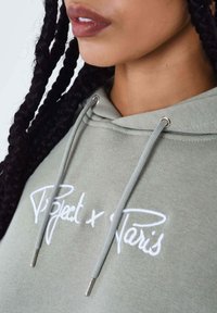 Olijfgroene hoodie met witte geborduurde tekst "Project x Paris." Voorzien van een trekkoord met zilveren aglets en een zachte stof.