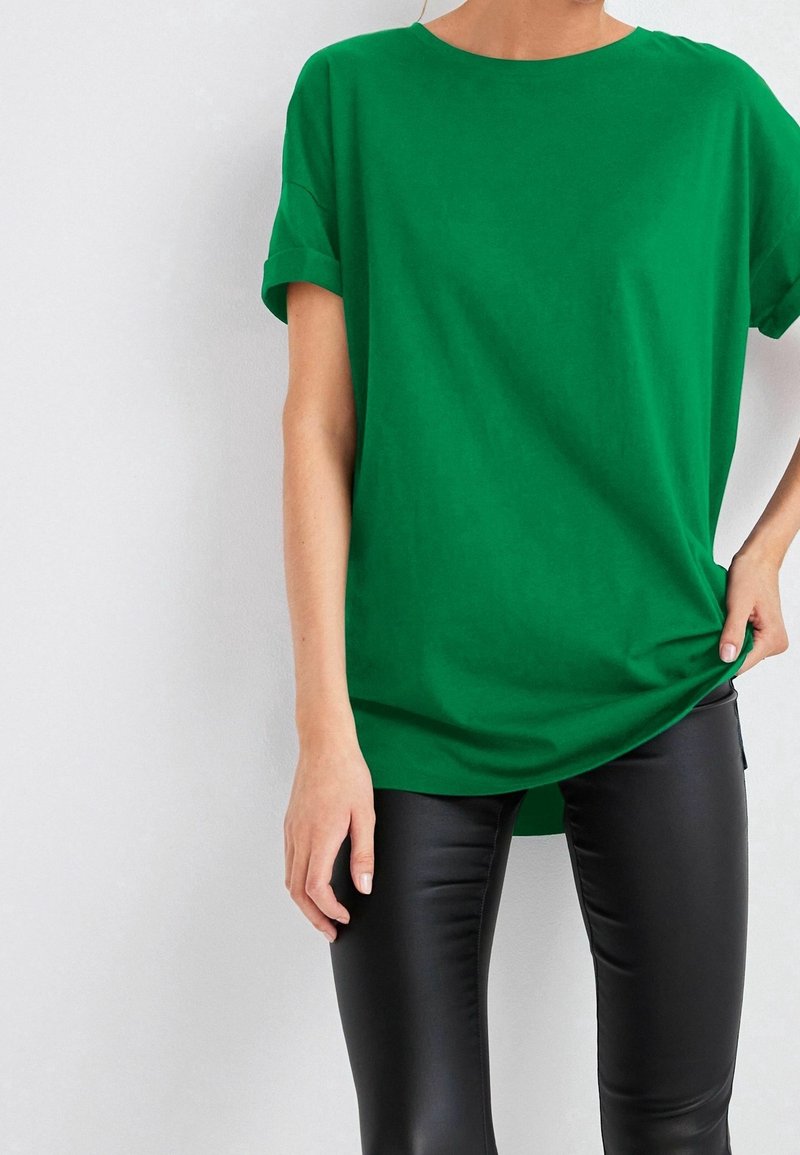 Femme portant un haut vert ample à manches courtes et un pantalon en cuir noir moulant, debout contre un mur blanc uni.