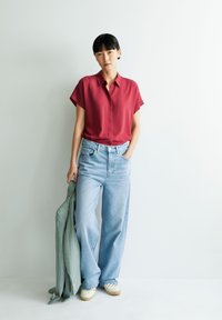 Rode blouse met korte mouwen, lichtblauwe wijde jeans, houdt een lichtgroene jas vast en crèmekleurige espadrilles, tegen een witte achtergrond.