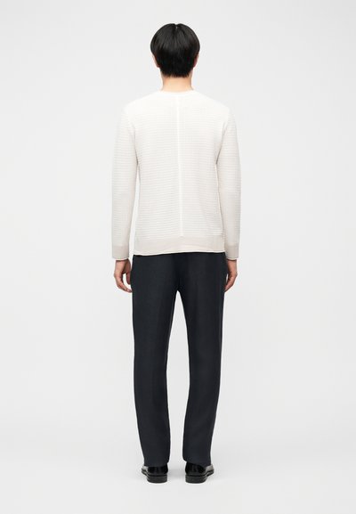 Άτομο με κοντά μαύρα μαλλιά που φοράει ένα άσπρο υφασμάτινο πουλόβερ μακρυμάνικο και μαύρο παντελόνι, στέκεται γυρισμένο προς τα πίσω μπροστά σε απλό φόντο.