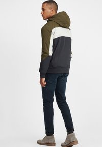 Hooded jacket with olive green, white, and black panels; features elastic cuffs and hem; worn with dark jeans and light gray boots.