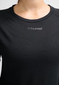 Close-up van een persoon die een zwart Hummel sportshirt draagt met een ronde hals en het merklogo op de borst.