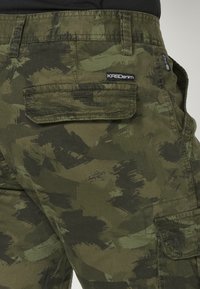 Vihreät ja tummanruskeat kamouflage cargo-housut, joissa on läppätaskut ja ommeltu "KRS Denim" -brändilogo takataskussa.