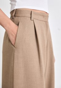 Beige geplooide broek vervaardigd van gestructureerde stof, met een tailleband met riemlussen en zijzakken, getoond met hand in de zak.