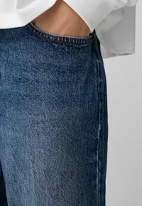 Roka v picah modrih jeans hlač, oblečen v belo majico z dolgimi rokavi, pred enobarvno svetlo sivo ozadje.