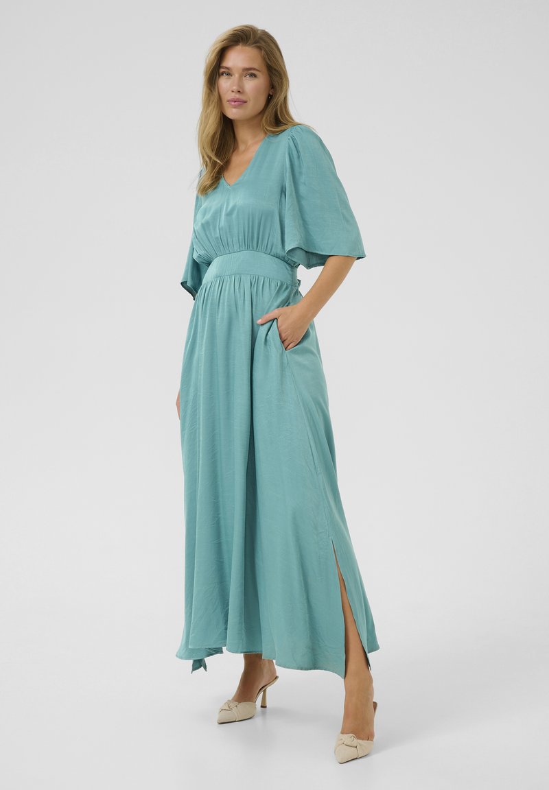 A long, light teal dress with a V-neck, short wide sleeves, cinched waist, side pockets, and slits on each side. Smooth fabric texture.