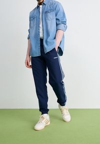 Ljusblå denimskjorta med två bröstfickor, vit t-shirt under, marinblå träningsoverallbyxor med vita sidostycken och krämfärgade sneakers.