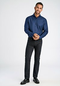 Donkerblauwe overhemd met knopen van glad stof, gecombineerd met zwarte slim fit broek en zwarte leren schoenen. Model staat met de handen bijeen.