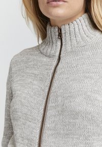 Maglione lavorato a maglia in grigio chiaro con colletto alto e zip bronzo. La texture è morbida e a coste, sottolineando un design casual.