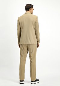 Completo beige con vestibilità su misura, caratterizzato da una giacca con spacco singolo e pantaloni a gamba dritta. Indossato con sneaker nere, texture in tessuto liscia.