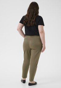 Olijfgroene broek met een slim fit en enkel lengte, gecombineerd met een zwart shirt met korte mouwen, gezien vanaf de achterkant met platte zwarte schoenen.