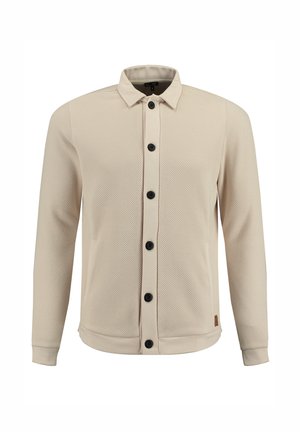 Chemise à manches longues de couleur beige clair en tissu maillé texturé, avec une fermeture à boutons à l'avant et des boutons noirs. Elle présente un col et des poignets élastiques.