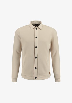 Camicia maniche lunghe color beige chiaro in tessuto a rete testurizzato, con chiusura frontale a bottoni neri. Presenta un colletto e polsini elastici.